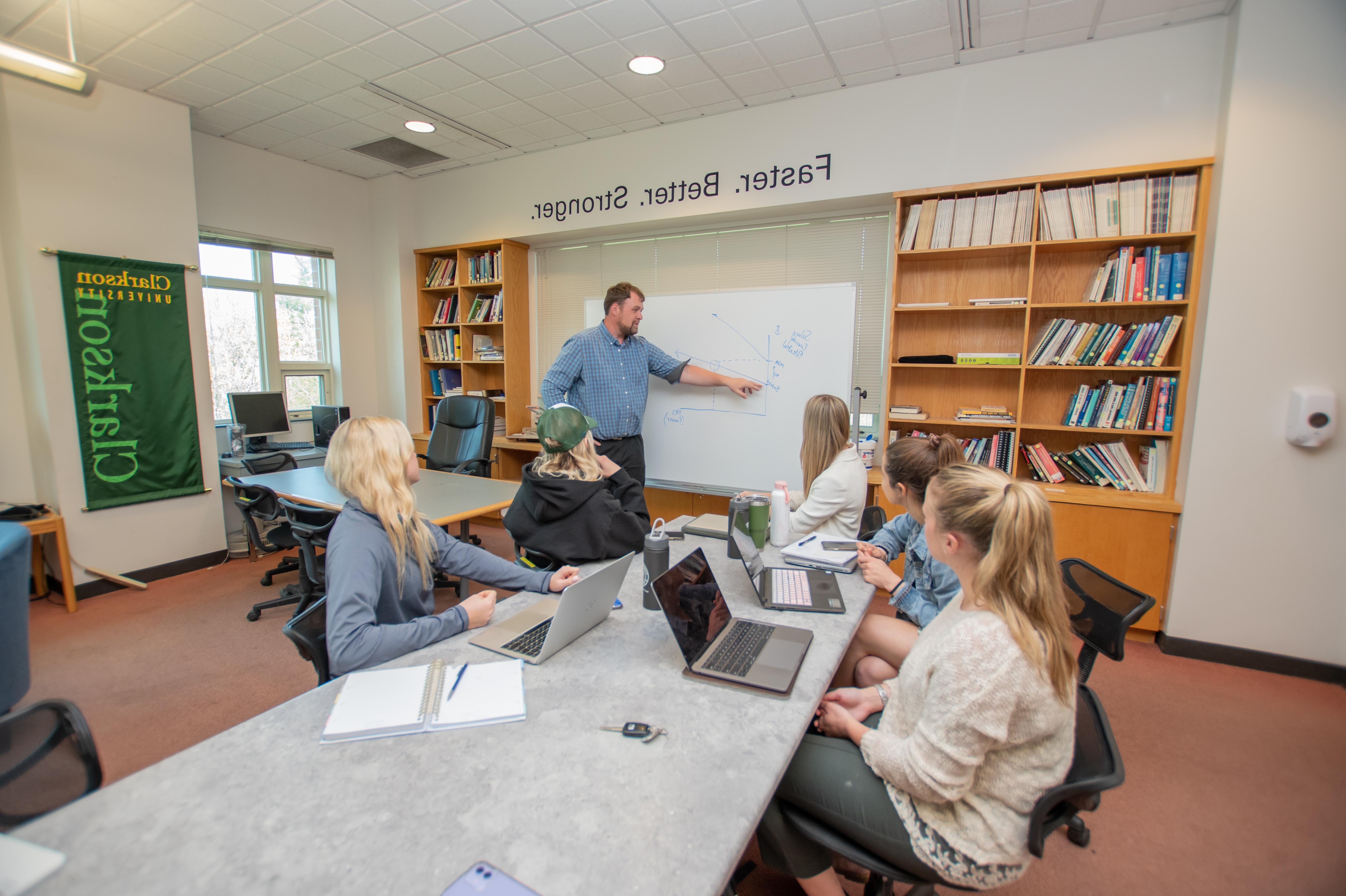 three students sit at a table with their laptops open as a professor points to a white board depicting a line graph with upward projections.